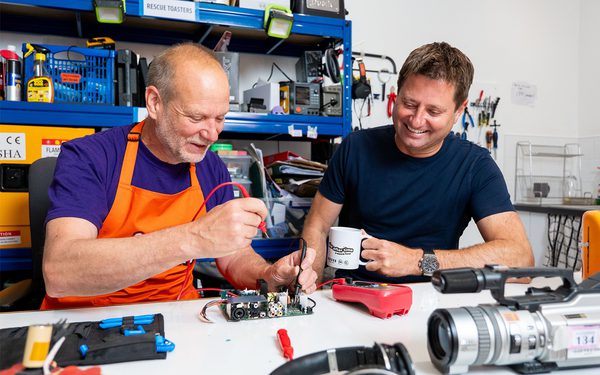 George Clarke is sat in a repair workshop watching a man repair an electronic circuit board.