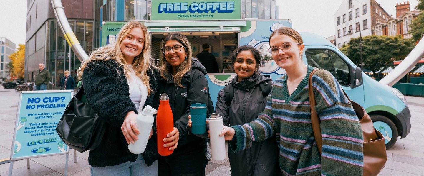 Four young women stand in front of a colourful van, with a sign saying 'Free coffee when you Bring Your Own Cup'. They are smiling and holding their reusable cups and water bottles towards the camera.
