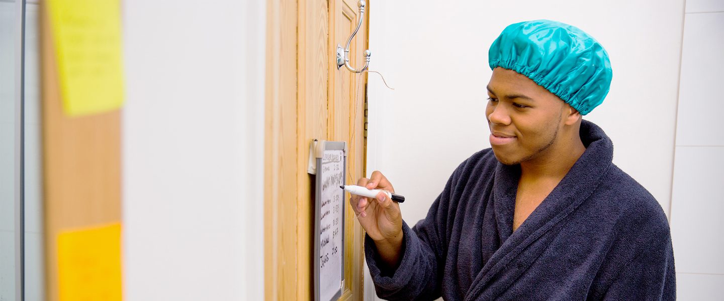 Person with blue shower cap and robe marking off a checklist on a whiteboard on the back of a door.