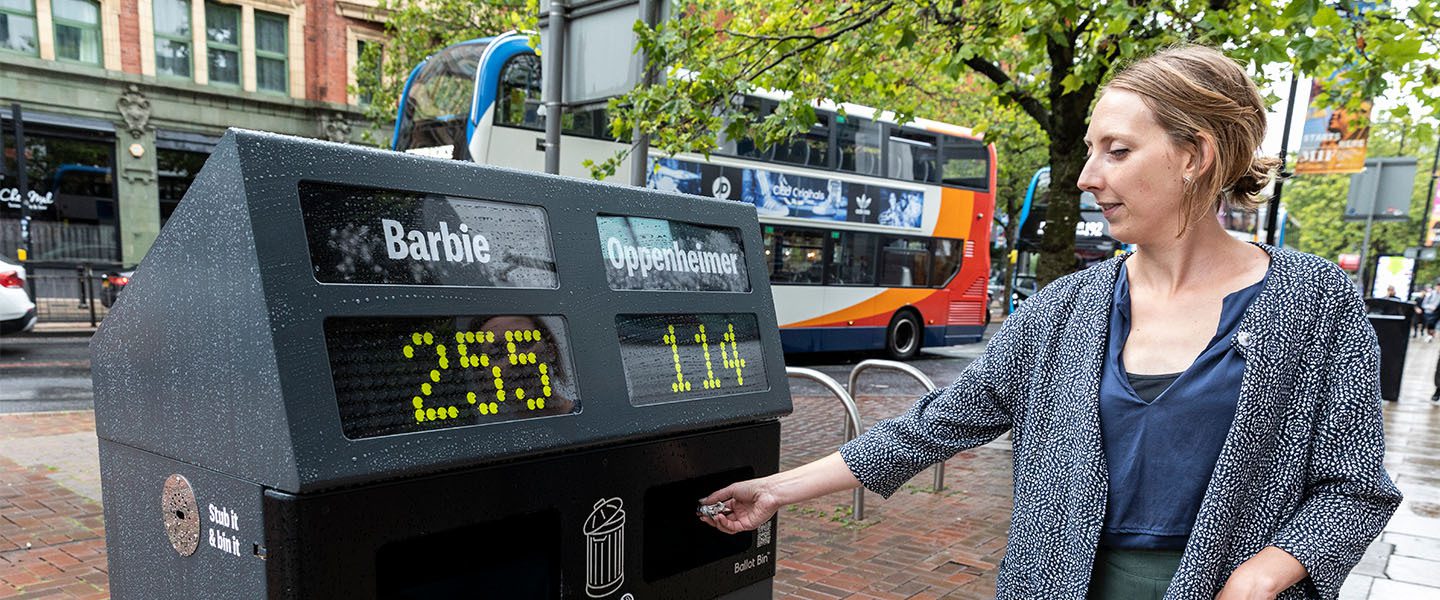 Someone dropping rubbish into a large ballot bin with two voting options for "Barbie" and "Oppenheimer".