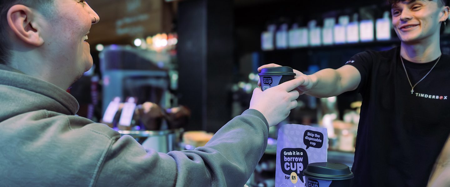 Barista handing a Borrow Cup to a customer over the counter. They are both smiling at each other. On the counter there is a strut card advertising Borrow Cup, a brand new returnable cup scheme for Glasgow, available at over 40 locations.