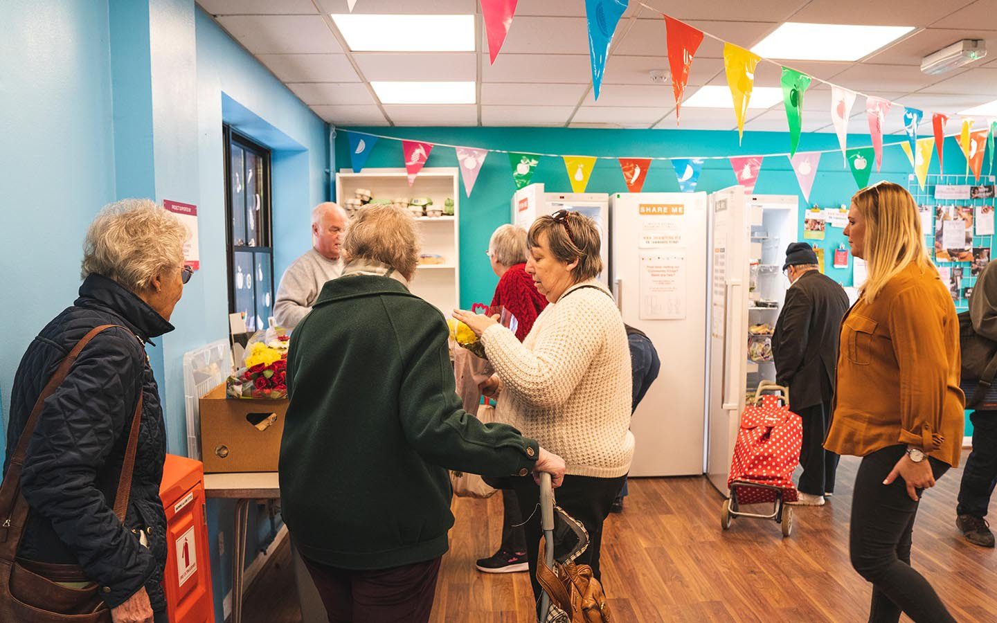 A group of people walking around a Community Fridge pantry with colourful banners hung from the ceiling.