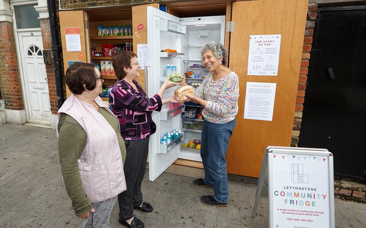 Someone handing another person a loaf of bread and some eggs from an outdoor Community Fridge.