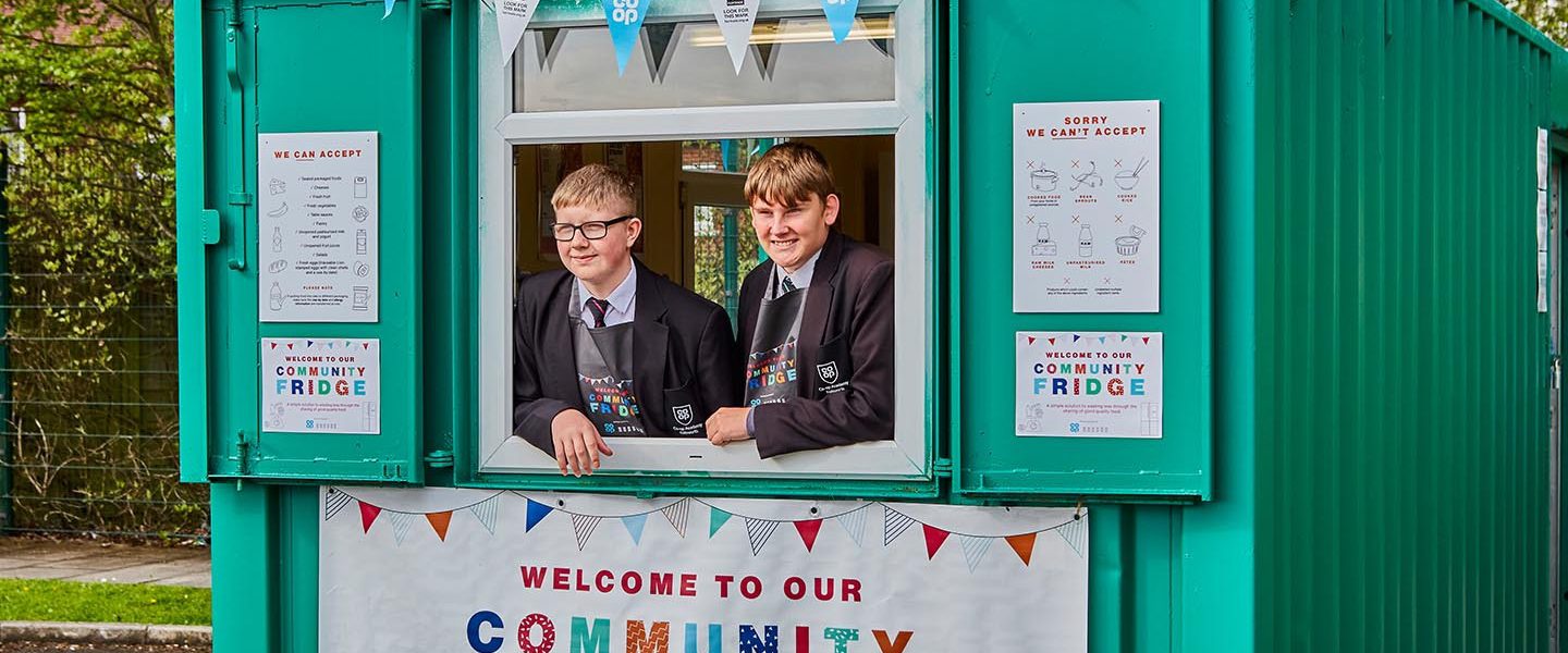 Two schoolchildren framed in the hatch of a green storage unit, which has been decorated with Community Fridge banners and branding.