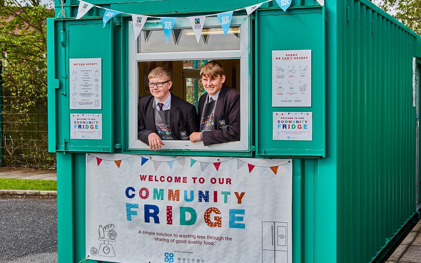 Two schoolchildren framed in the hatch of a green storage unit, which has been decorated with Community Fridge banners and branding.