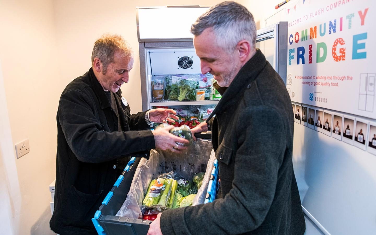 Two people taking food out of a community fridge and placing in a hamper.