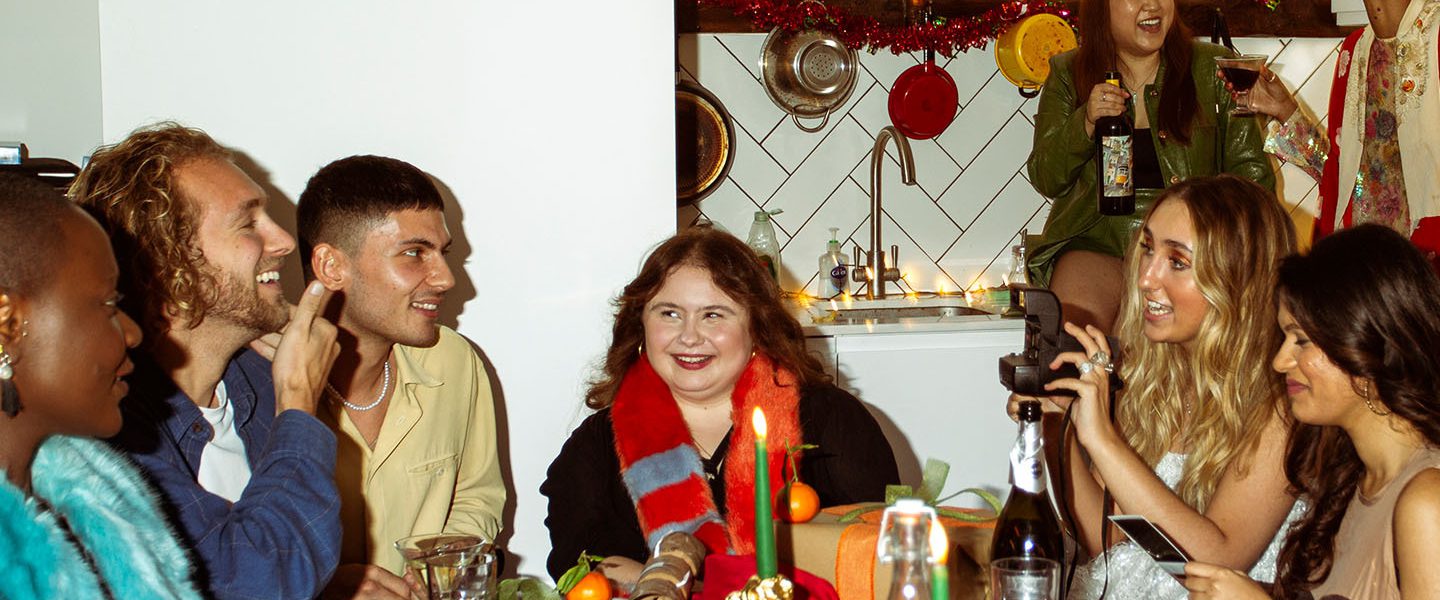 A diverse group of people in their twenties or thirties laughing and chatting around a festive table with candles and oranges, in a kitchen decorated with fairy lights.