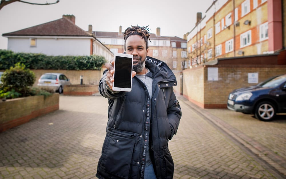 A young black man standing in the middle of a street holding their new smartphone from Community Calling to the camera.