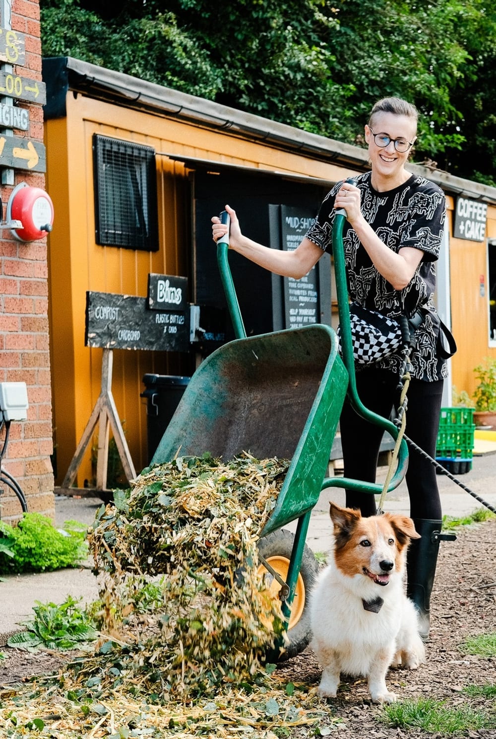 Woman lifting up a green wheelbarrow to dump organic matter onto a garden patch. They are smiling and looking down at their dog on a lead that is tethered to their waist.