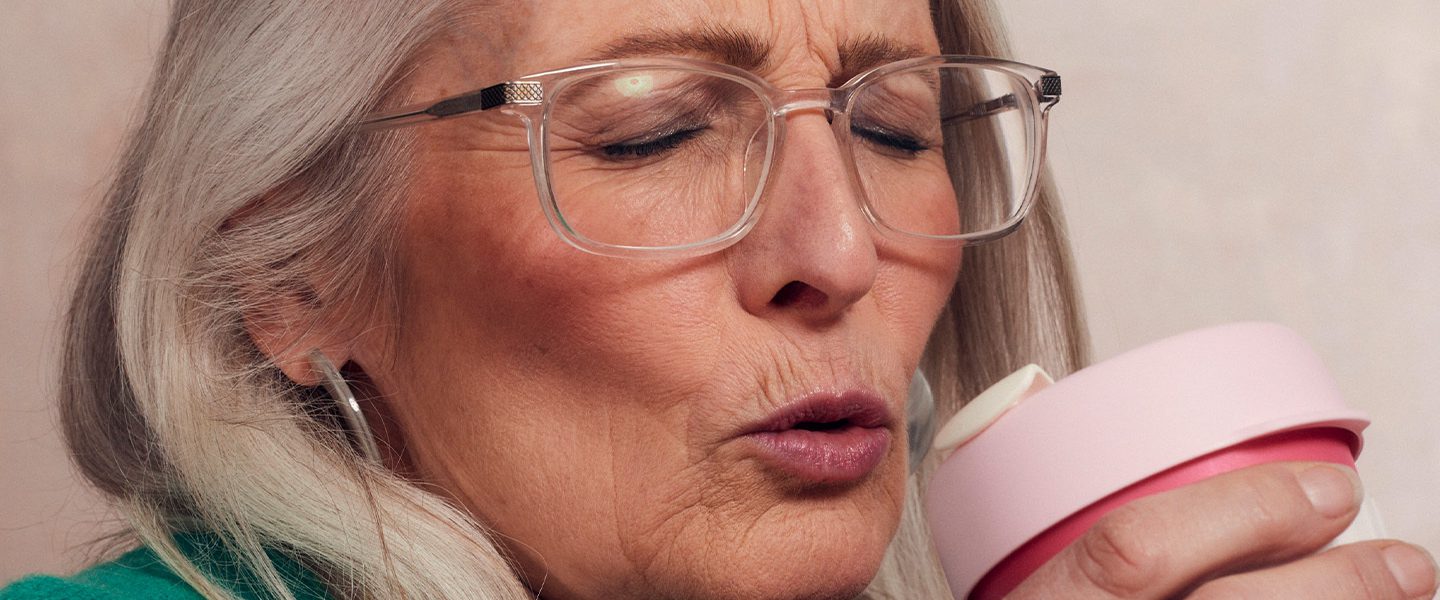 Close up shot of glamorous senior woman with glasses closing her eyes and pursing her lips into an 'o' shape as she brings her pink reusable coffee cup up to her face.