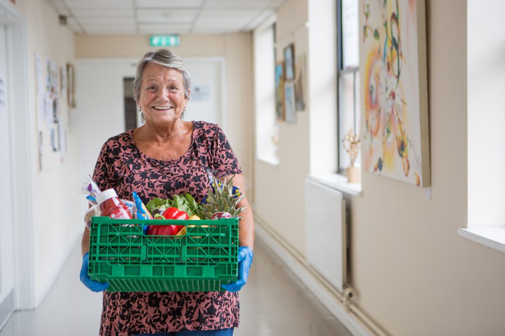 A smiling woman carrying a green crate full of food, walking down a corridor towards the camera.