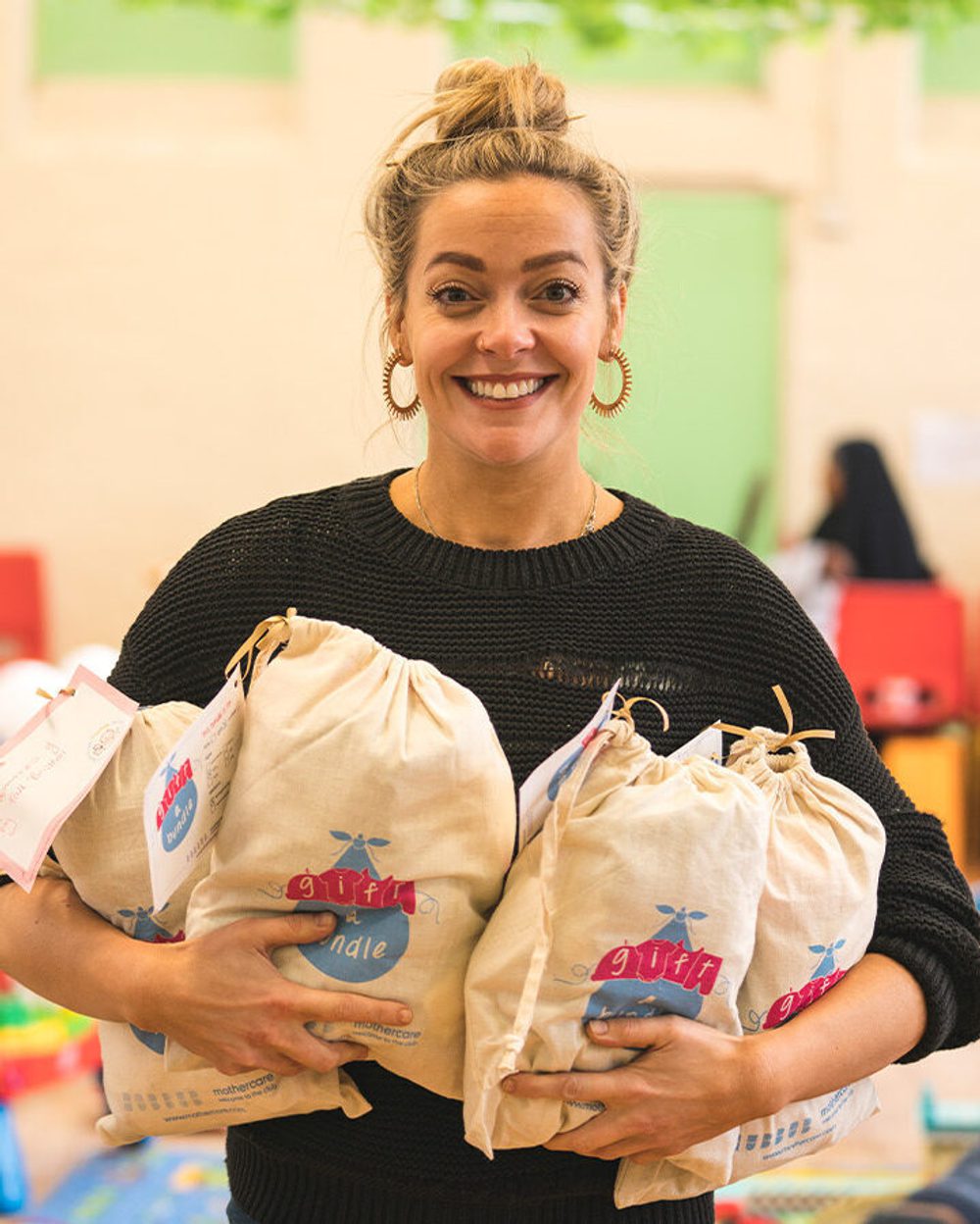 A blonde woman holding four Gift a Bundle bags containing pre-loved baby clothes ready to be gifted to new parents.