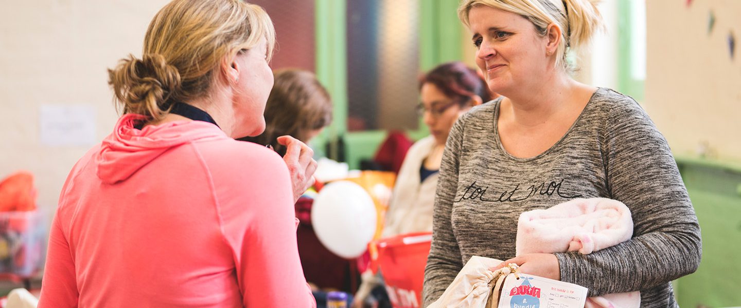 Two white woman holding Gift a Bundle bags containing pre-loved baby clothes speaking to each other.
