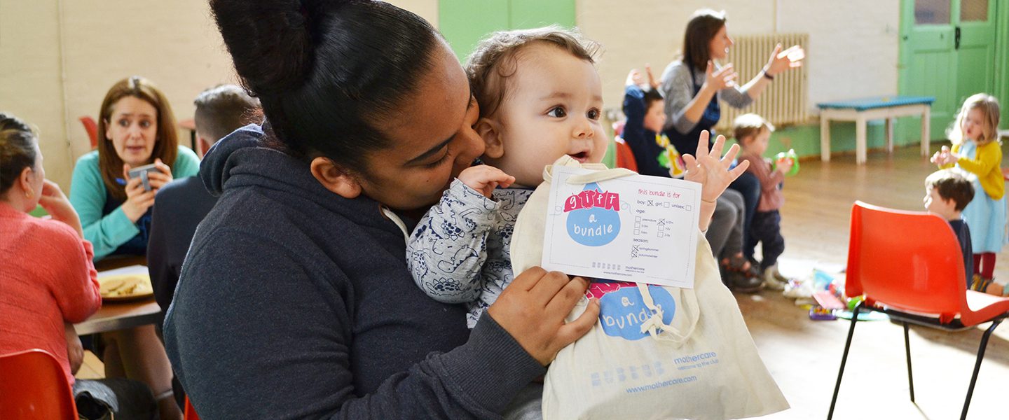 A black mother holding her baby in a community hall with other mother. She is also holding a Gift a Bundle bag containing pre-loved baby clothes.