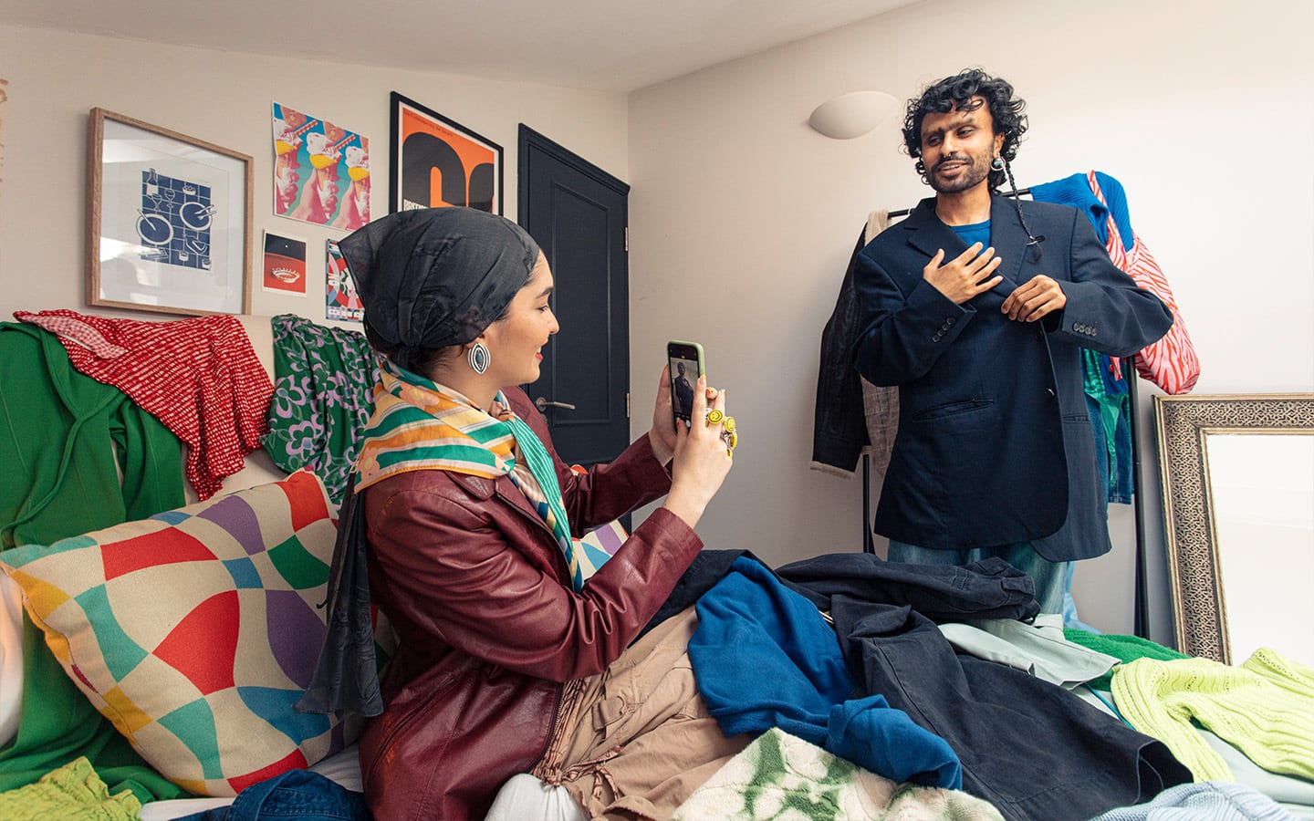A woman with a scarf on her head is sitting on a bed covered in clothes she is taking a photo of her male friend who is trying on clothes.