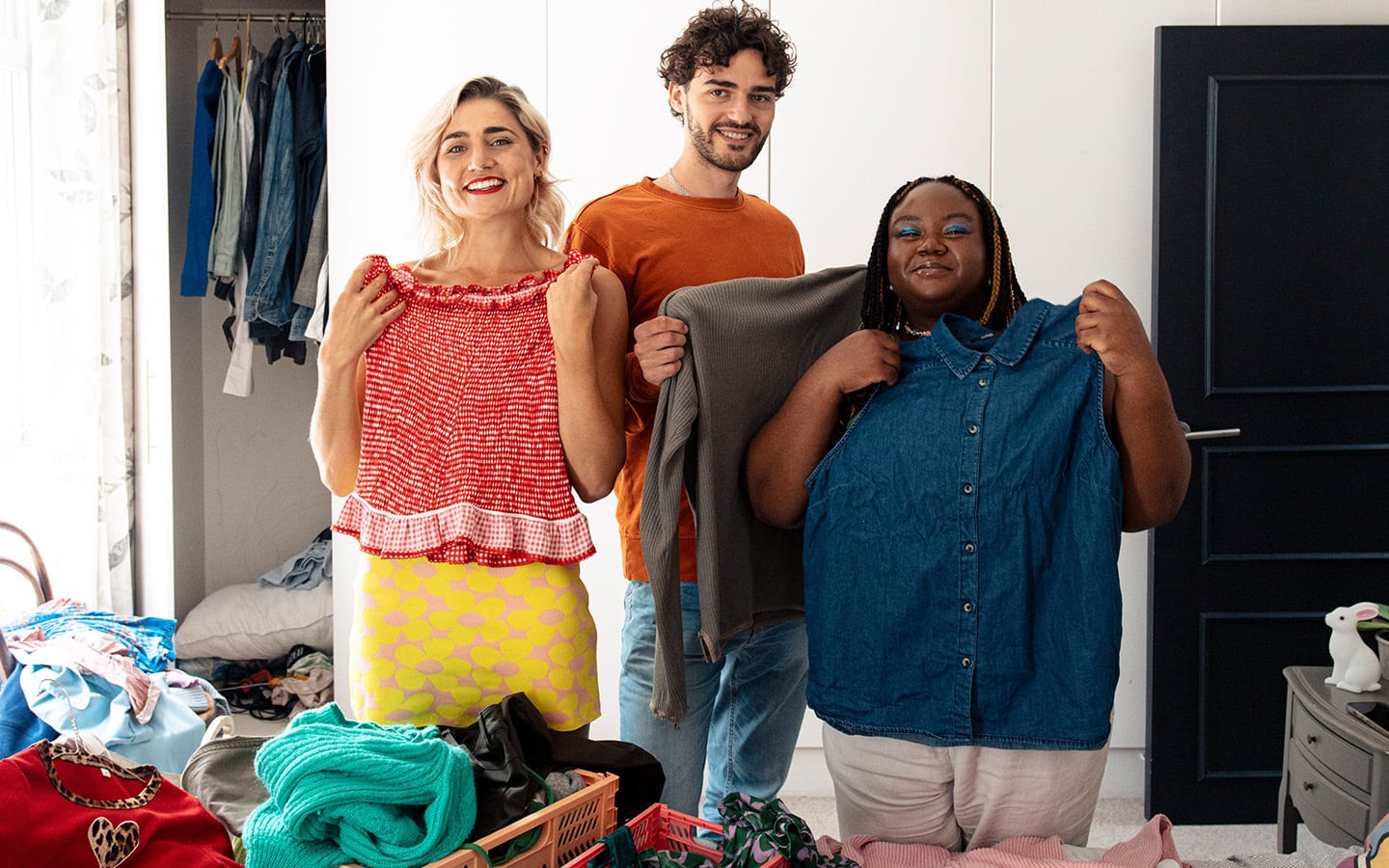 Three friends in a bedroom are holding up different items of clothing and smiling at the camera.