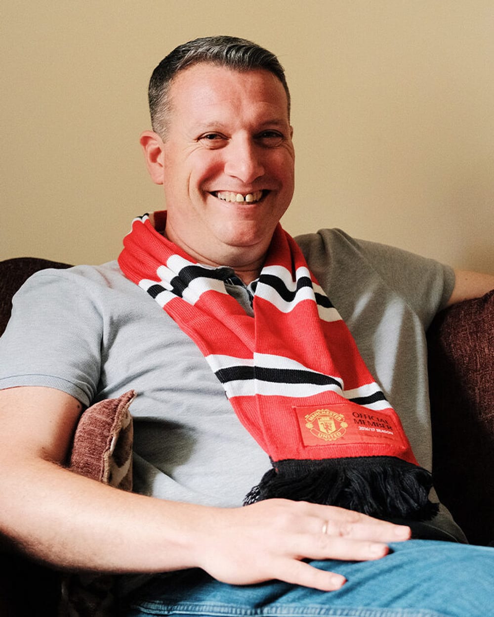 A man sat on his sofa smiling, he is wearing a Manchester United football club scarf.
