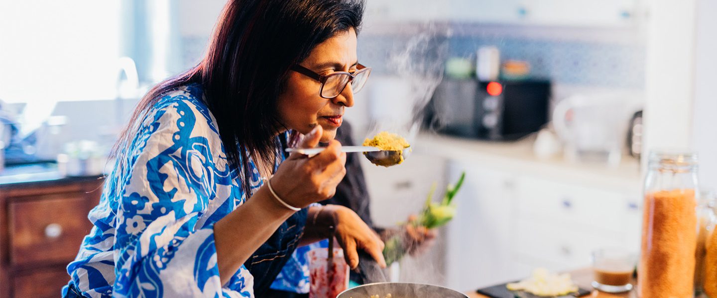 Someone stood over a pan of hot food in their kitchen, smelling a spoonful of food from the pan.