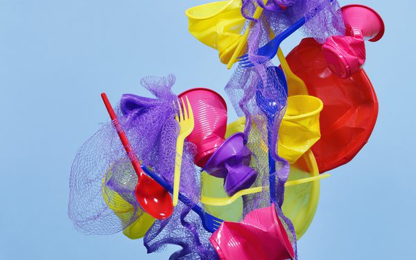 Purple, yellow and red plastic items such a cutlery and cups are cascading towards the ground against a blue background.