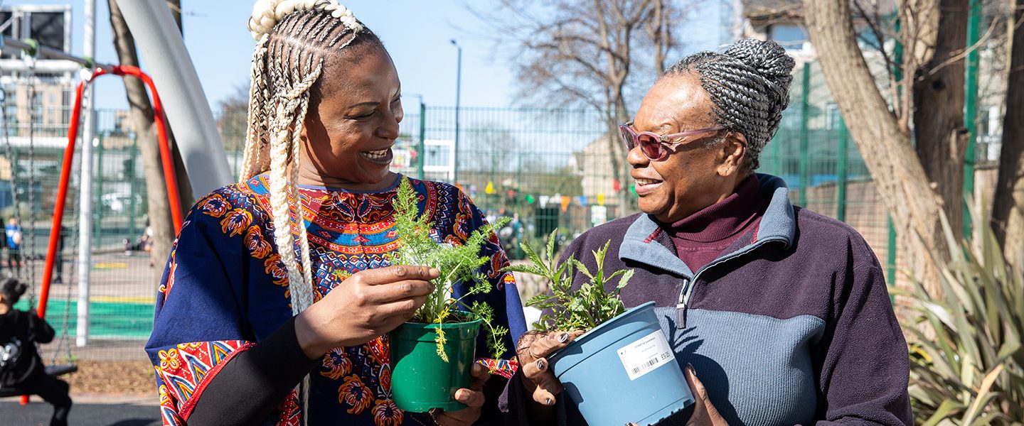 Two black woman stood next to each other on a children's play area, each holding potted plants and smiling at one another.