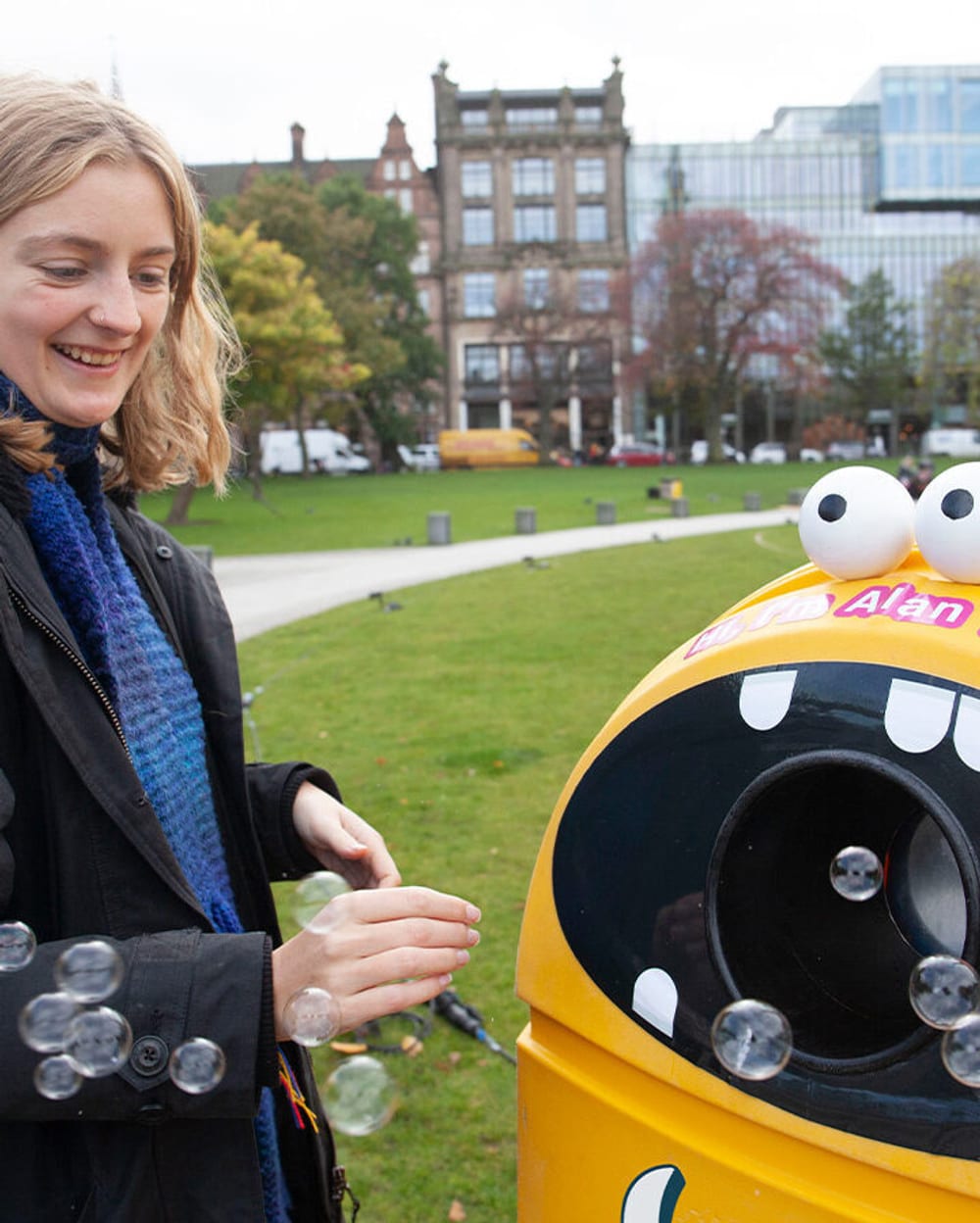 A young woman putting recycling into a cartoon monster bin that blows bubbles when you 'feed' it recycling.