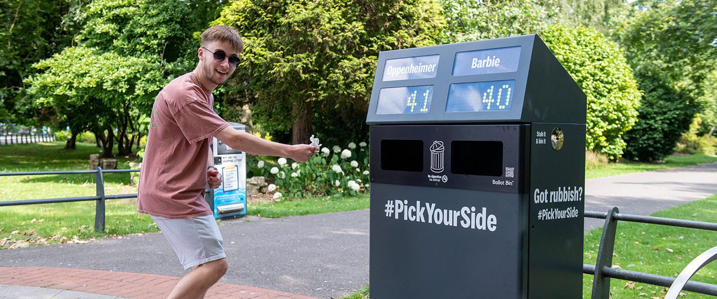 A young man excitedly putting into the 'big ballot bin' and voting with their litter. The voting options are Barbie and Oppenheimer.
