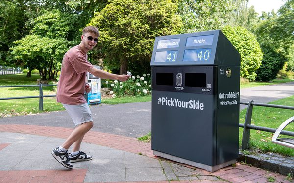 A young man excitedly putting into the 'big ballot bin' and voting with their litter. The voting options are Barbie and Oppenheimer.