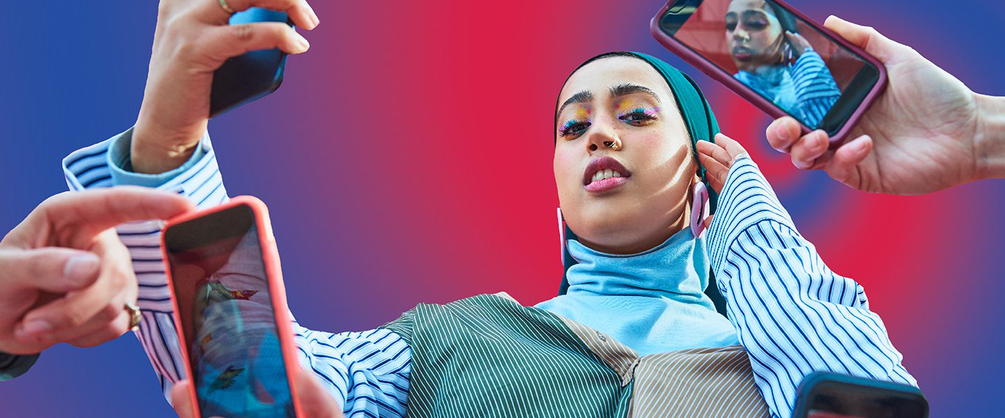 A young woman with a headscarf and colourful eyeshadow is holding a phone to take a selfie. Around her are lots of different people holding phones taking her picture. The background is a graphic image of a purple and blue swirl.