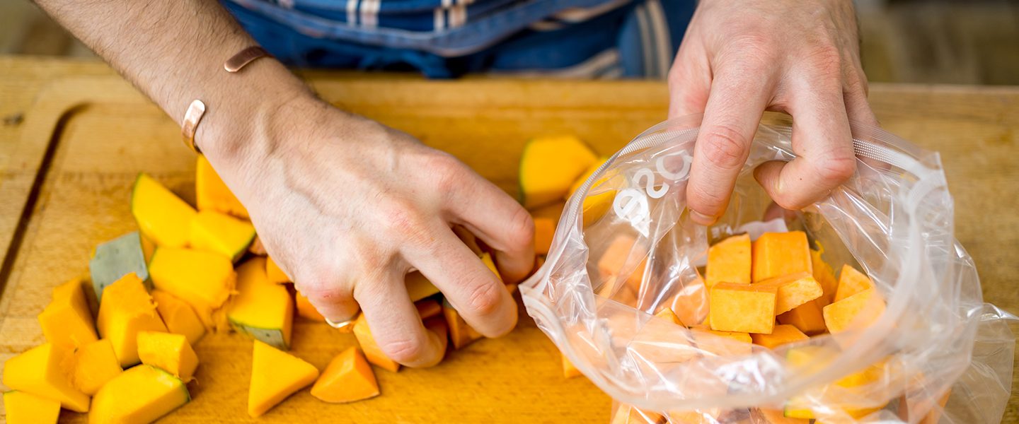 A man is picking up chopped pieces of squash from a chopping board and putting them in a freezer bag ready to be frozen.