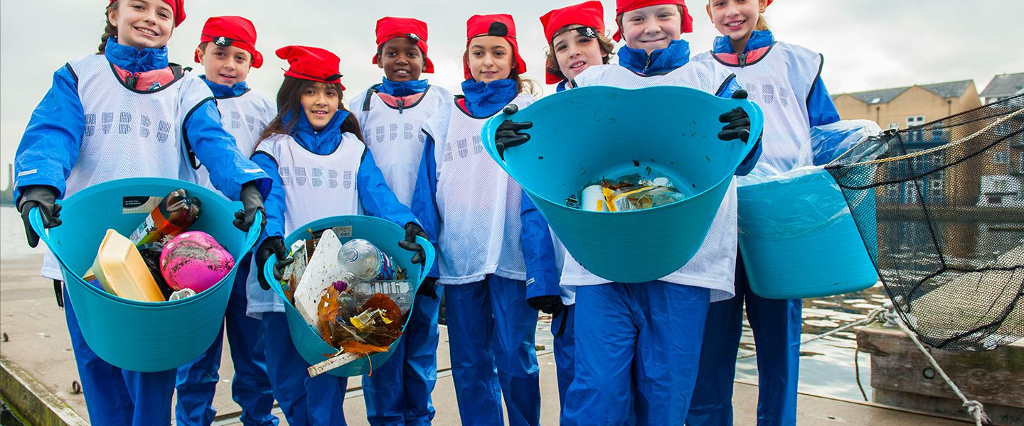 A group of school-aged children with red scarfs on their heads and blue waterproof outfits are stood smiling on a dock. They are holding buckets of litter that they have just 'fished' out of the river from their Plastic Fishing trip.