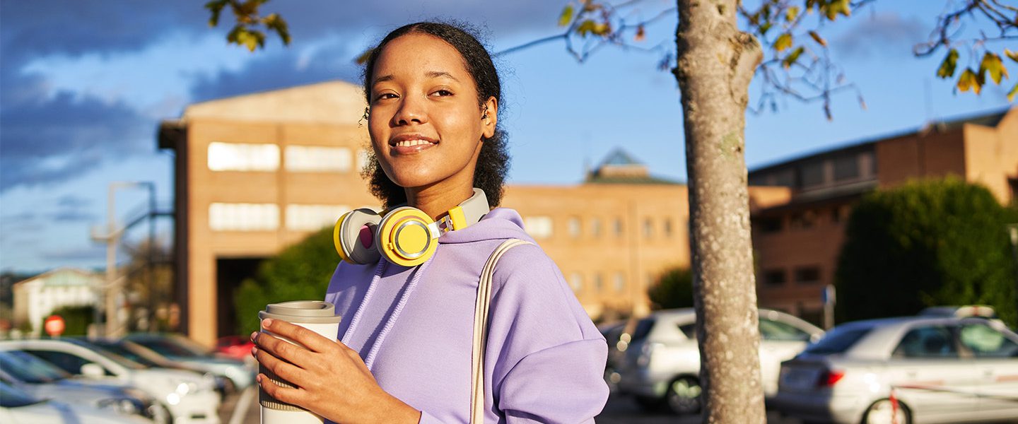 A young mixed-race woman in purple workout gear standing in a carpark holding a grey reusable coffee cup.