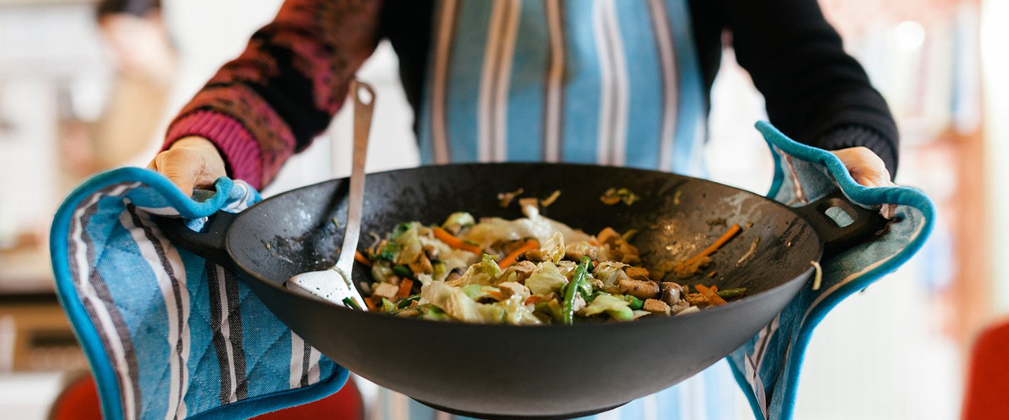 A woman wearing a blue striped apron is holding a wok with fried vegetables in it.