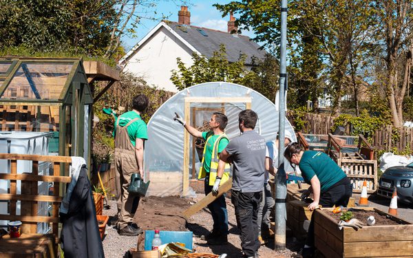 5 volunteers working on creating a community garden. The garden has sheds on the left, a polytunnel at back and a pile of pallets to the right.