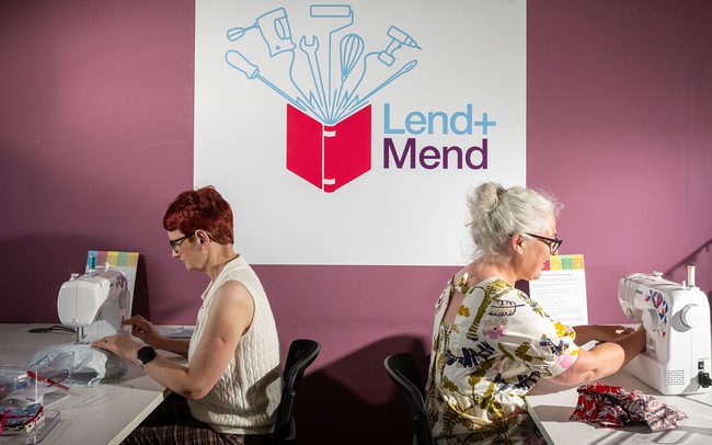 Two women sitting with their backs to each other and using sewing machines to mend garments. Behind them is the logo for 'Lend + Mend'.