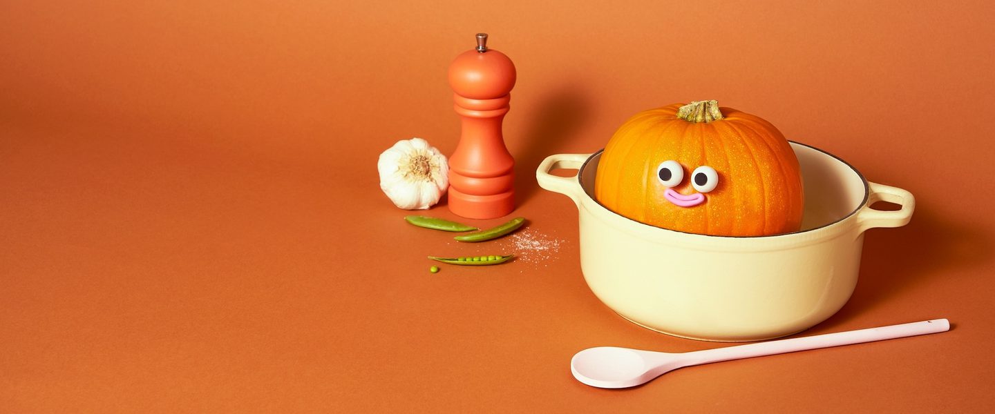 A decorated pumpkin with a cute face is photographed inside a dutch over cooking pot, with a spoon and salt shaker also on the table.