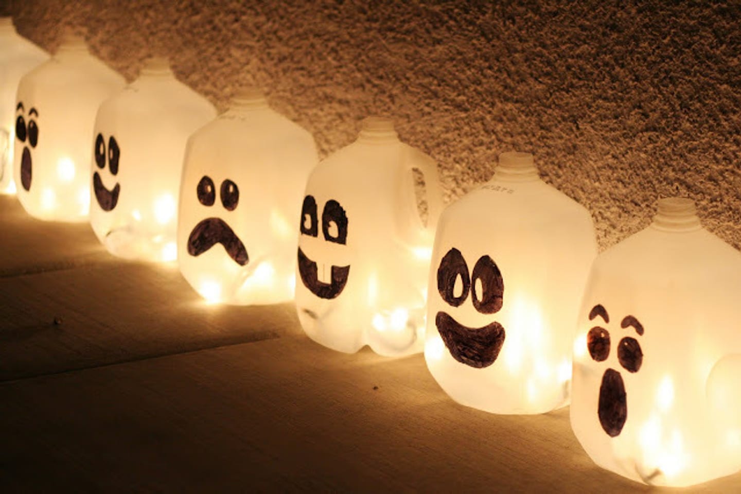 6 milk plastic milk bottles decorated with Halloween pumpkin-style faces. There are lights inside them lighting them up like a Halloween pumpkin.