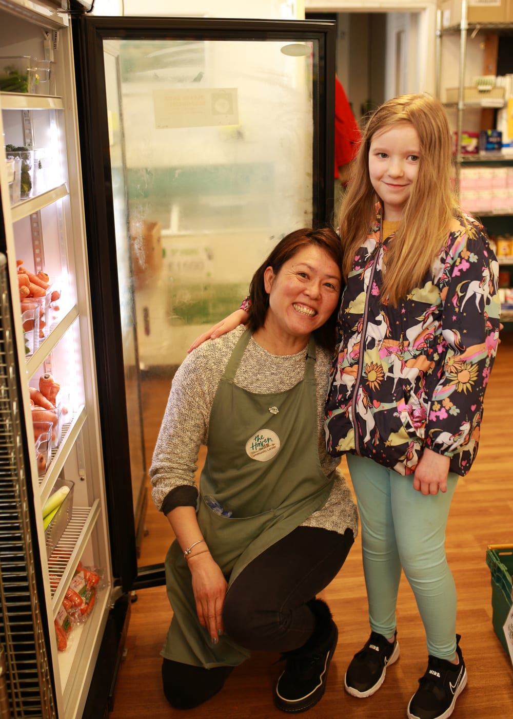 Volunteers at a community fridge. An adult is crouched down, smiling, in front of an open fridge. To her right, a young girl is standing and smiling at the camera.