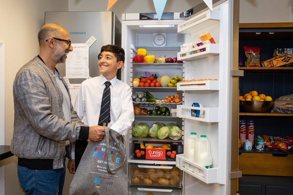 A father and son smiling to one another whilst stood in front of an open community fridge filled with food.