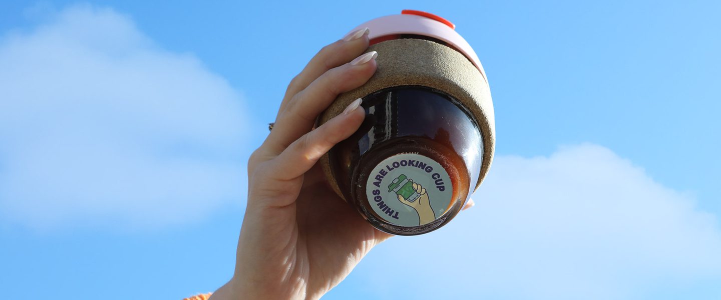 A hand reaches up to the sky, holding a glass reusable cup with coffee in it. Under the reusable cup, a light blue sticker reads 'things are looking cup'.