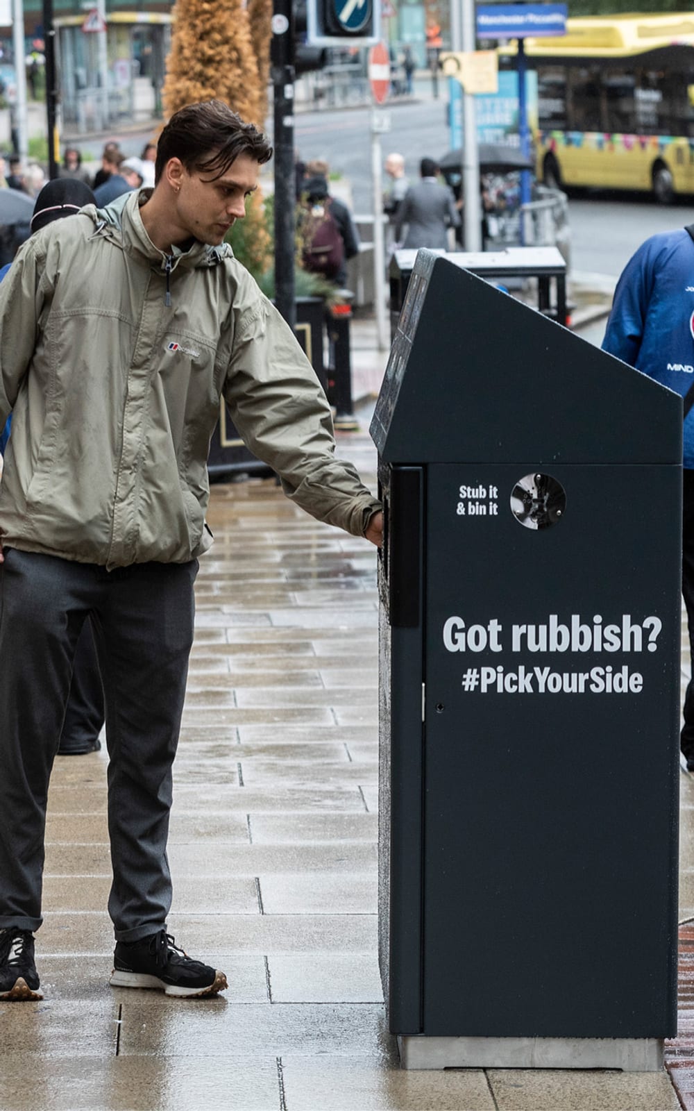 A young man placing is litter in a 'big ballot bin' in Manchester. The side of the bin reads 'Got rubbish? #PickYourSide'.
