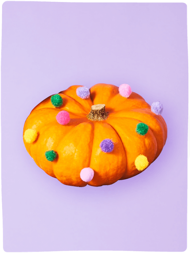 Flat pumpkin decorated with colourful pompoms sat against a light purple backdrop.