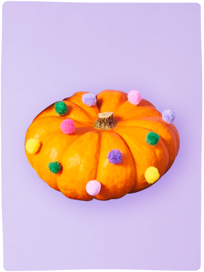 Flat pumpkin decorated with colourful pompoms sat against a light purple backdrop.