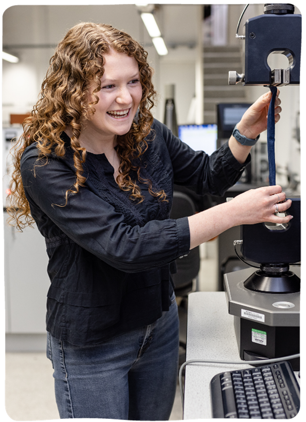 A young woman with curly hair is standing smiling while operating a machine in a lab.