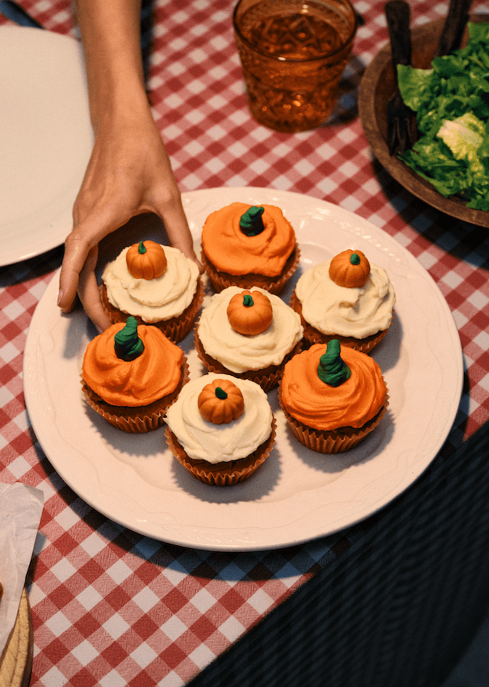 Top view of seven pumpkin themed cupcakes on a white plate, placed on a red and white gingham table cloth. From the top of the frame, a hand in reaching in the take one of the cupcakes.