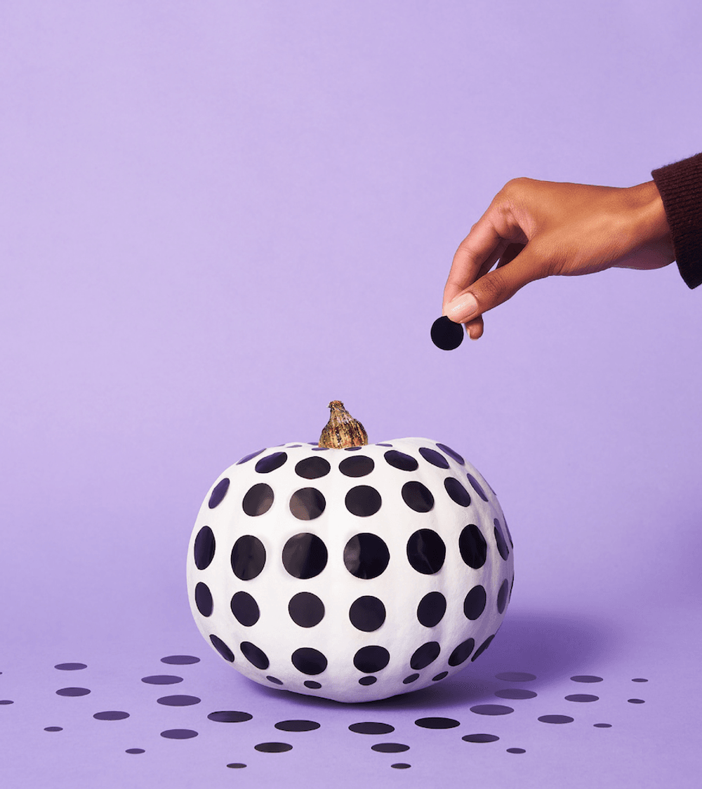 A painted white pumpkin decorated with black dots sat against a light purple backdrop. A person's hand is reaching into the frame to add a dot to the pumpkin. There are dots on the floor radiating out from the pumpkin.