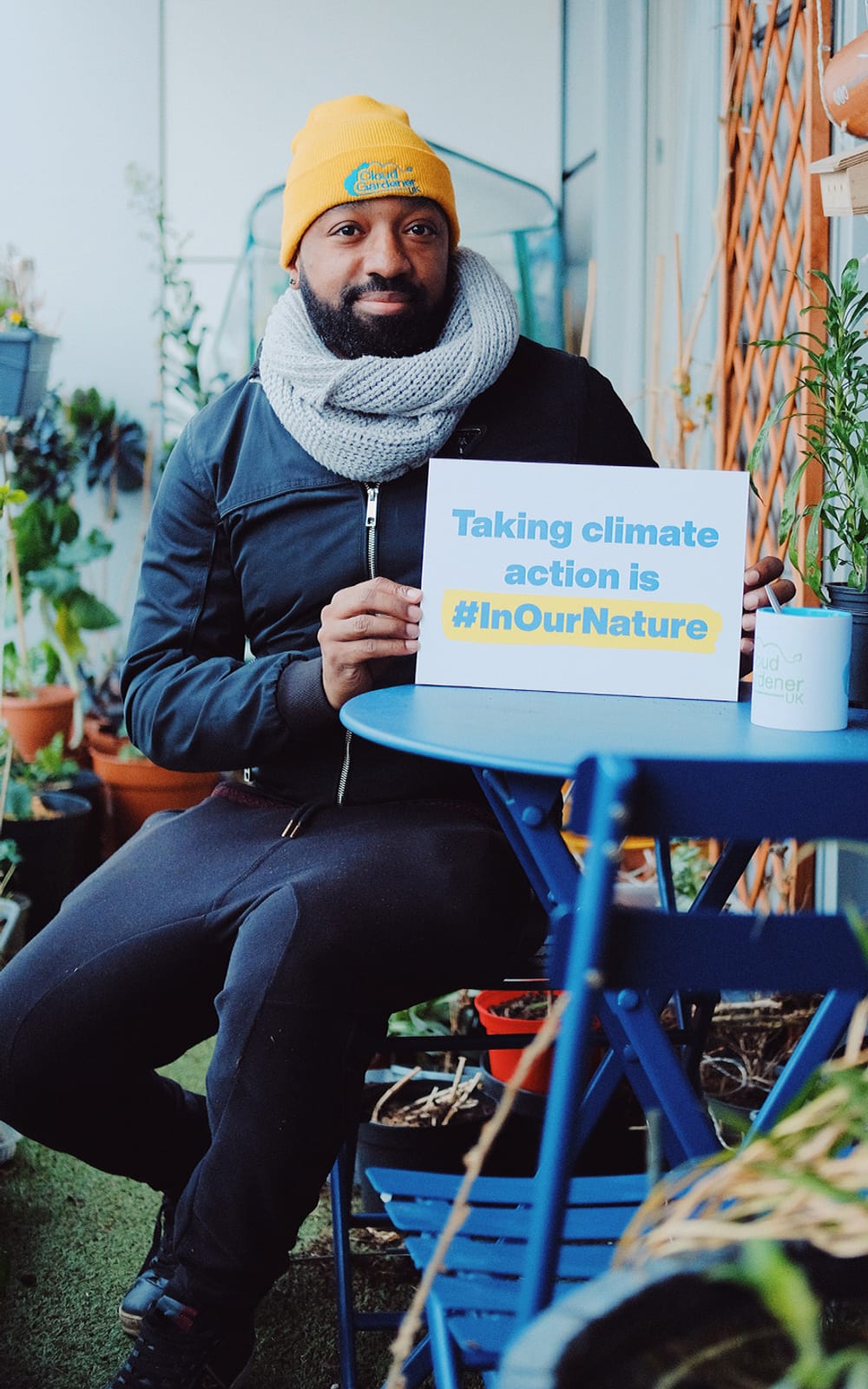 A black man sitting in a garden on blue patio furniture. He is holding a sign that says 'taking climate action is #InOurNature'.