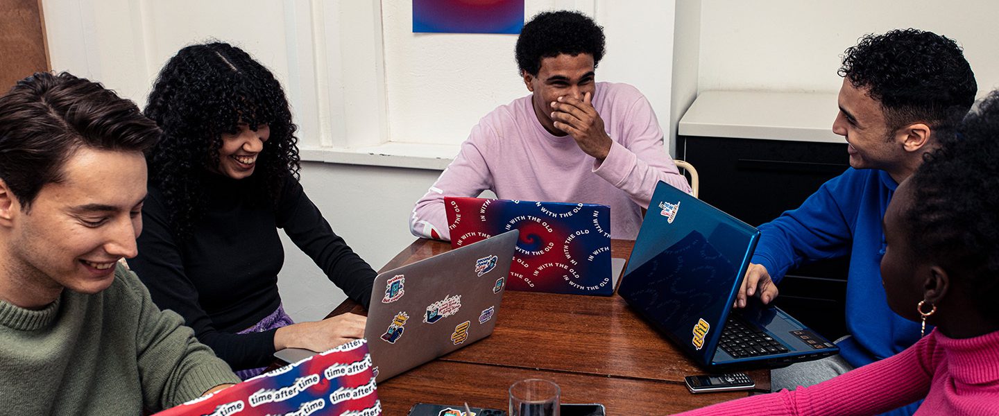 A group of students are sitting around a table laughing and talking. They have their laptops out that have been branded with Time After Time stickers and coverings.