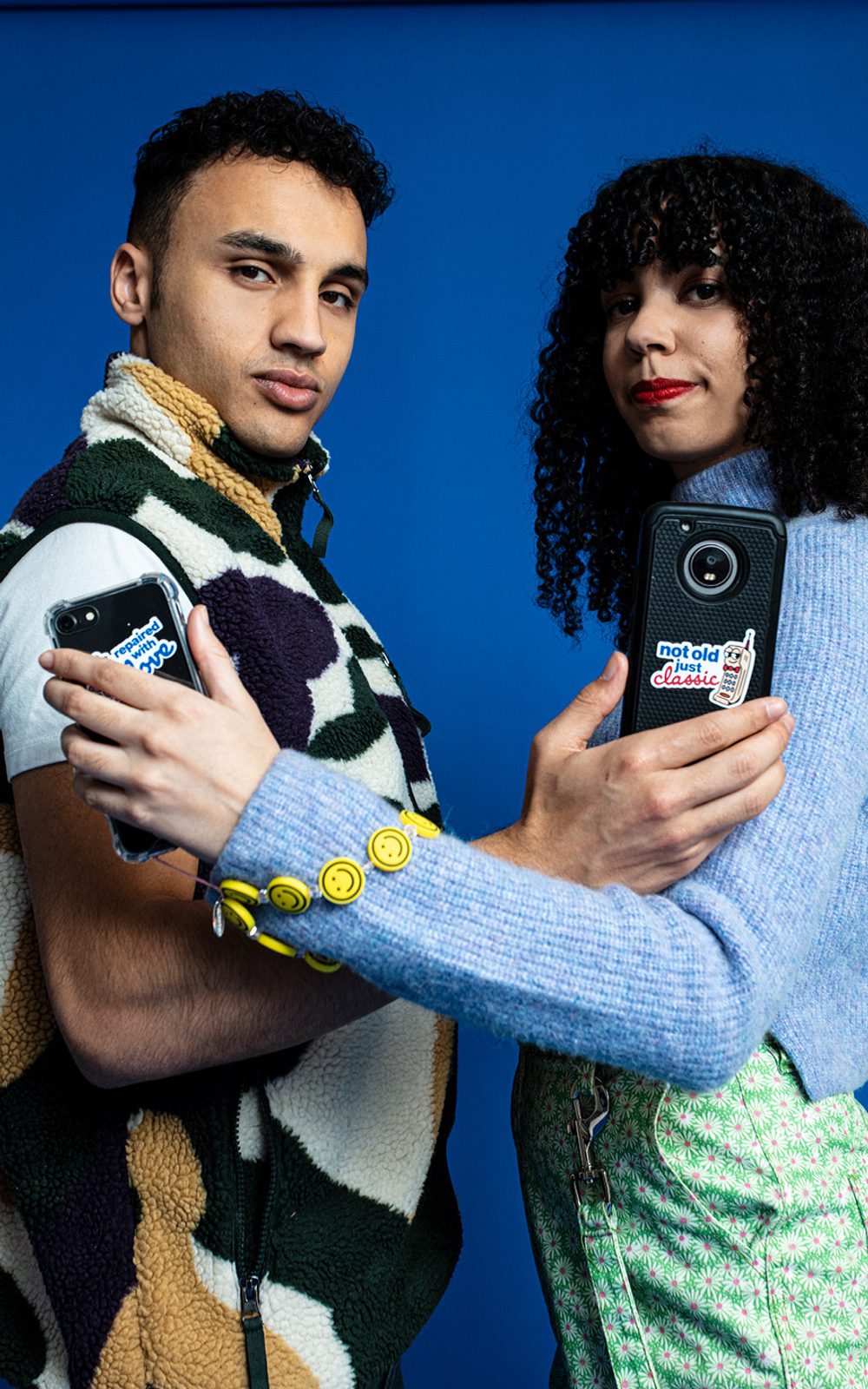 A young man and woman are standing opposite each other looking at the camera. They have their arms interlinked while holding up phones towards the camera.