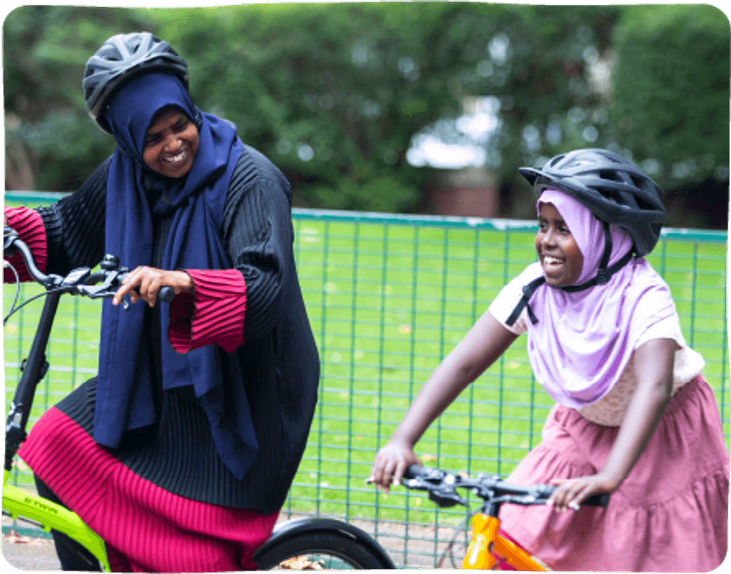 An adult and a child riding bicycles next to one another, with a large green space in the background.