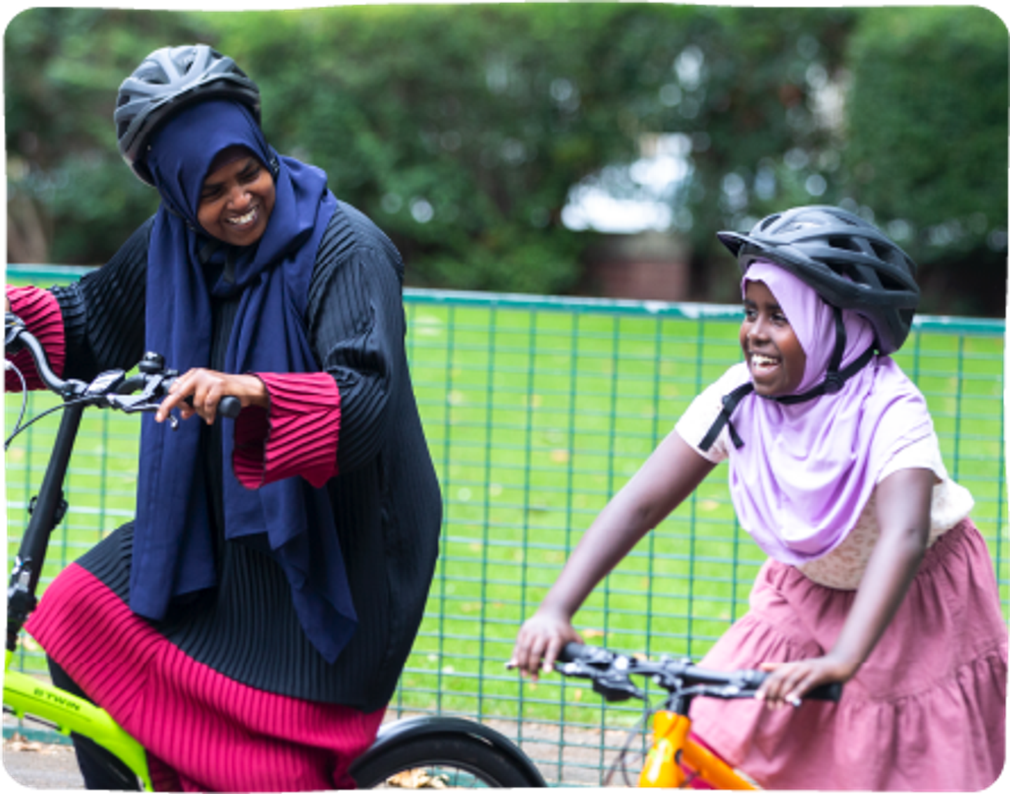 An adult and a child riding bicycles next to one another, with a large green space in the background.
